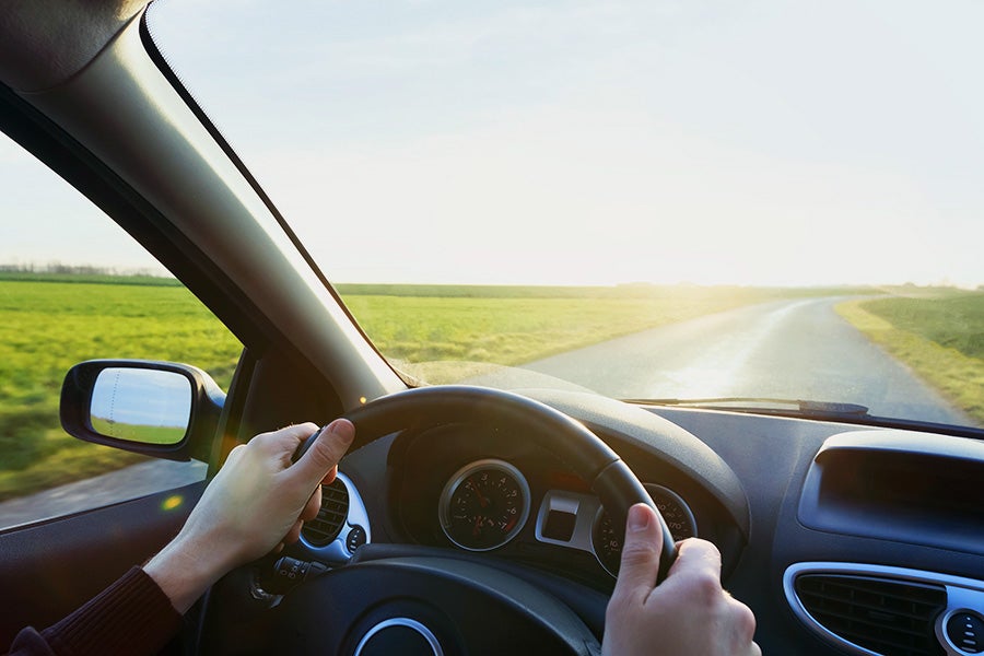 Driver's hands on the steering wheel with a view through the windshield of a rural road and green fields during a sunny day.